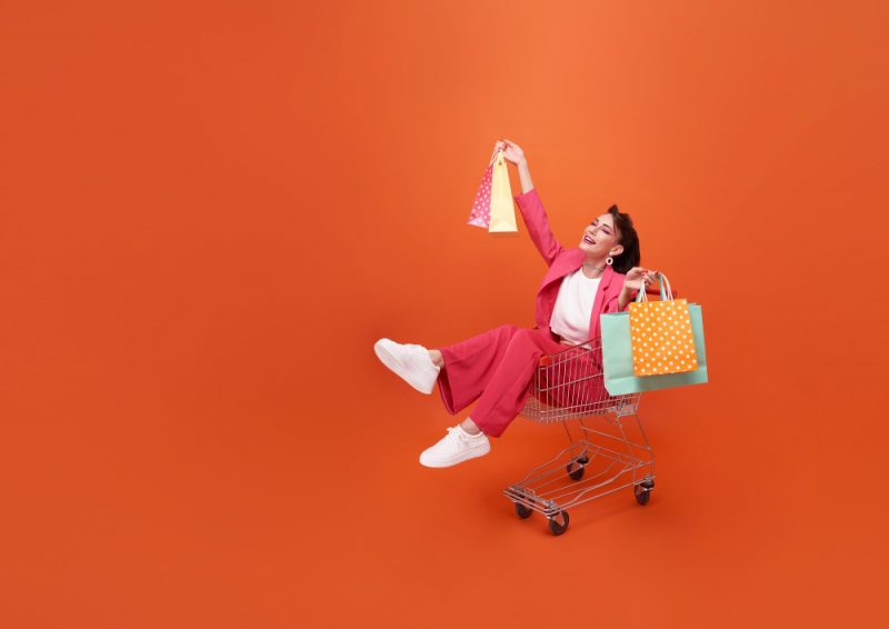 Woman in a pink suit sitting in a shopping cart, holding colorful shopping bags against a bright orange background, expressing joy and excitement from shopping