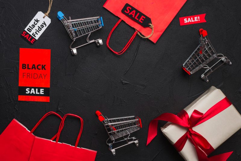 Mini shopping carts, sale tags, and a gift box with a red ribbon arranged on a dark background, highlighting Black Friday sale themes.